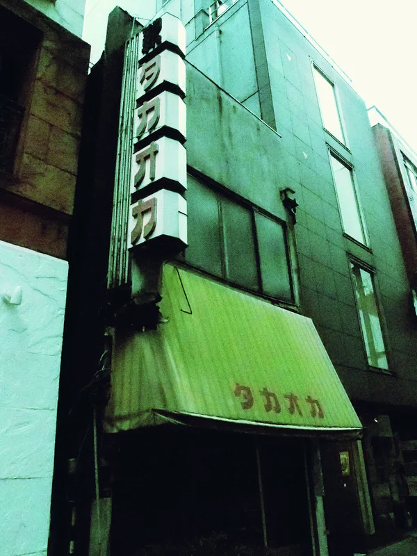 A tall green-toned facade has a vertical character sign and a pale green entrance awning.