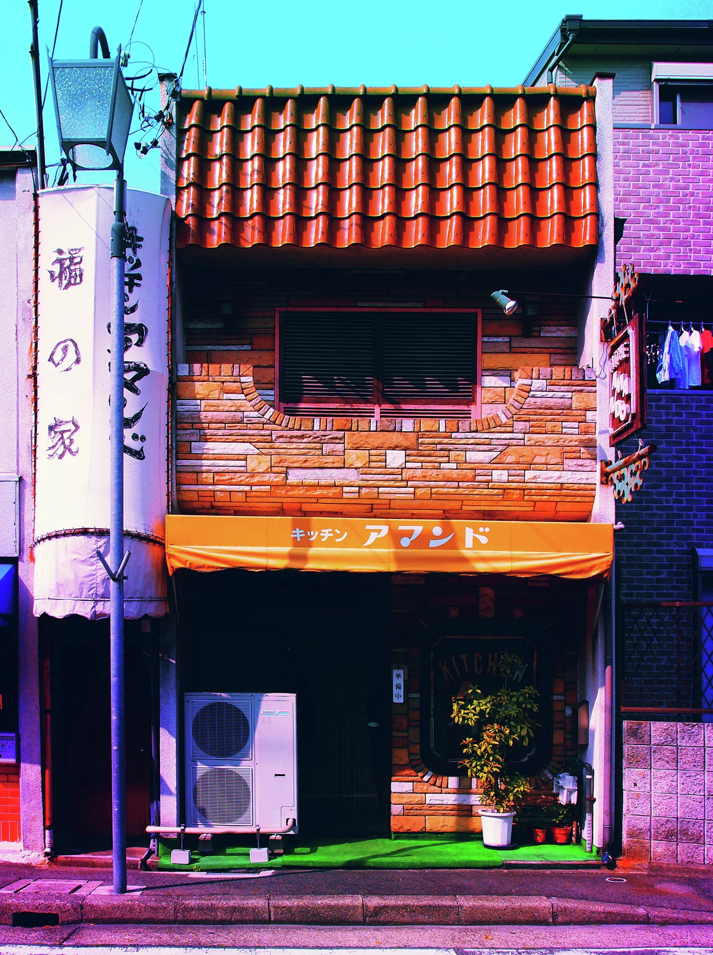 An orange tiled roof and awning top a narrow brick-fronted shop with potted plants.