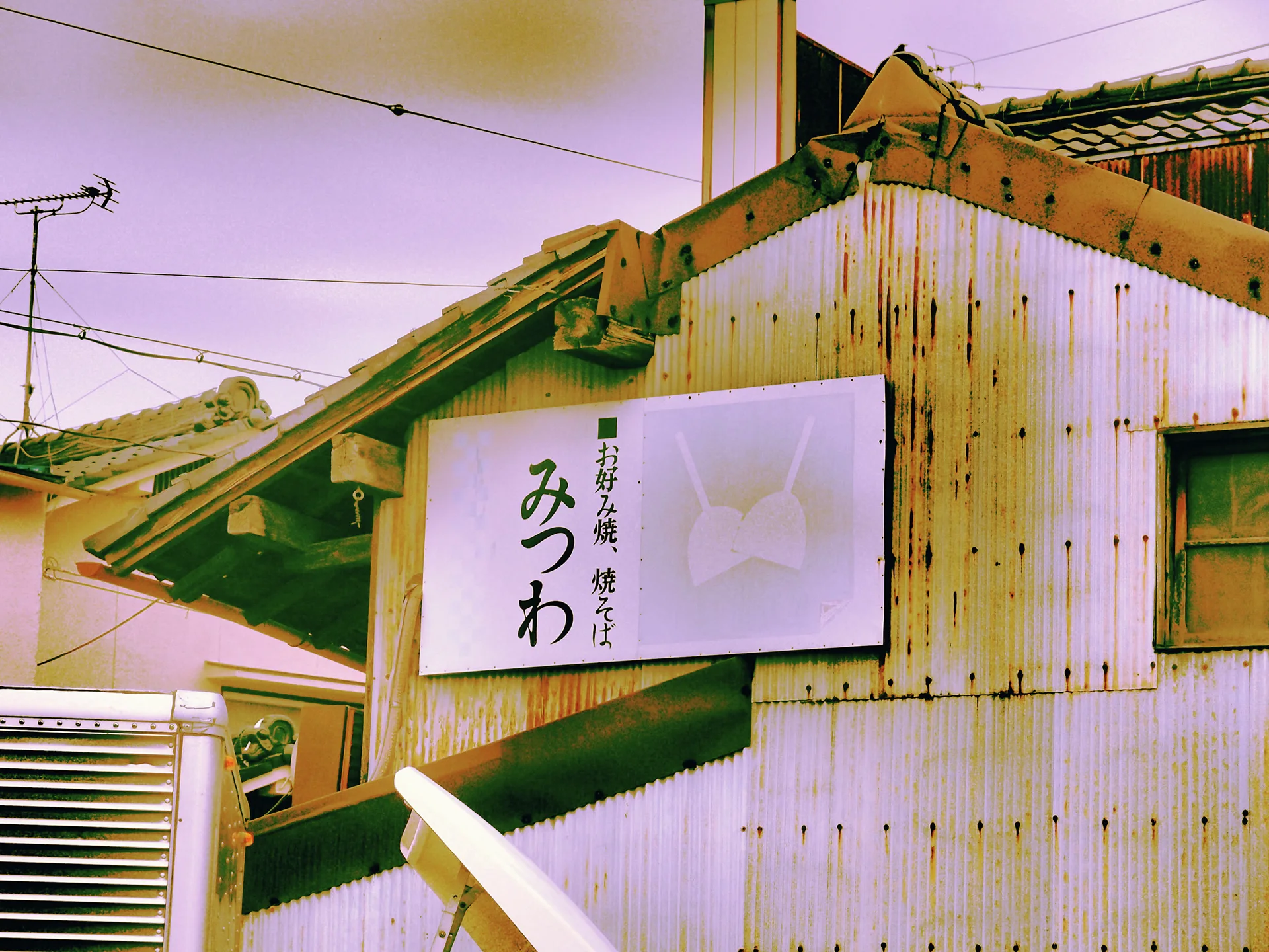 A white restaurant sign hangs on rust-streaked corrugated siding beneath a tiled roof.
