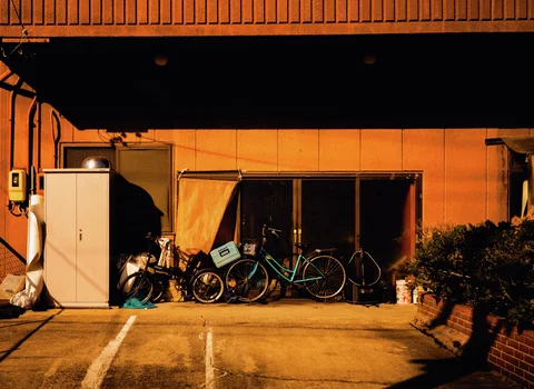 Several bicycles lean beneath a window on an orange-lit wall beside a tall pale cabinet.