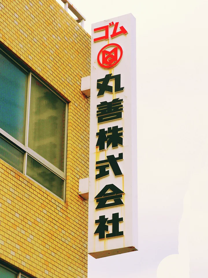 A tall white Japanese sign with a red emblem projects beside yellow brickwork and a broad window.
