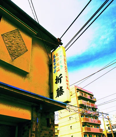 A vertical Japanese sign projects from a yellow building beneath wires and blue sky beside taller apartments.