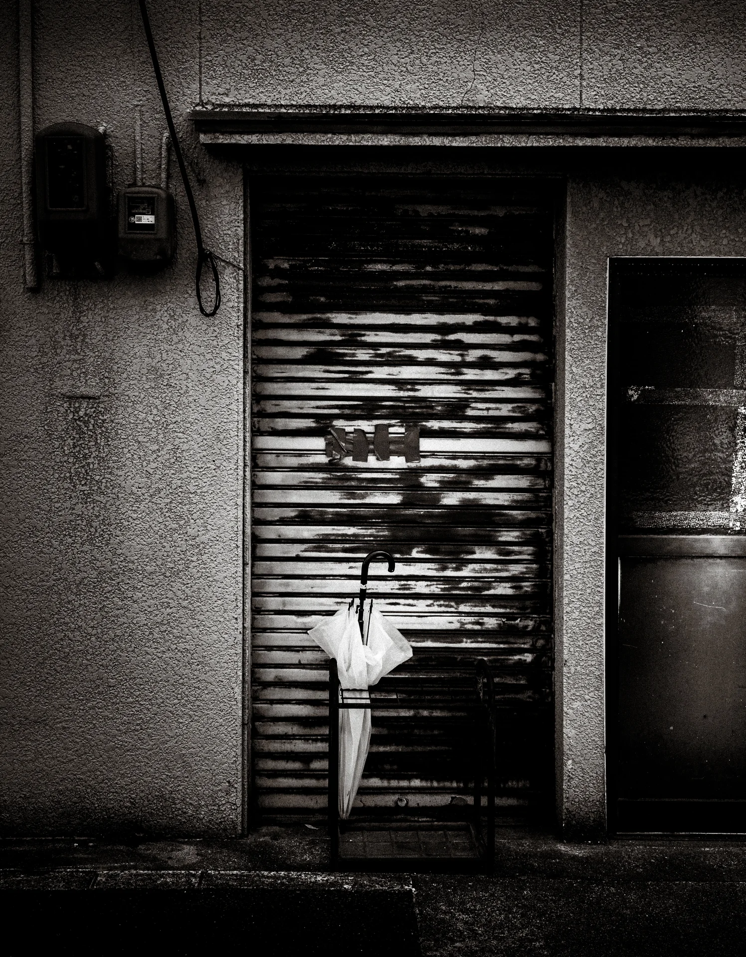 A folded umbrella hangs against a peeling metal shutter beside a dark doorway in black and white.