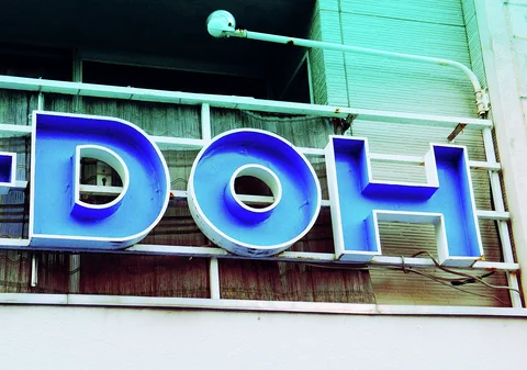 Large blue metal letters project from a white frame before textured windows and a pale corrugated wall.