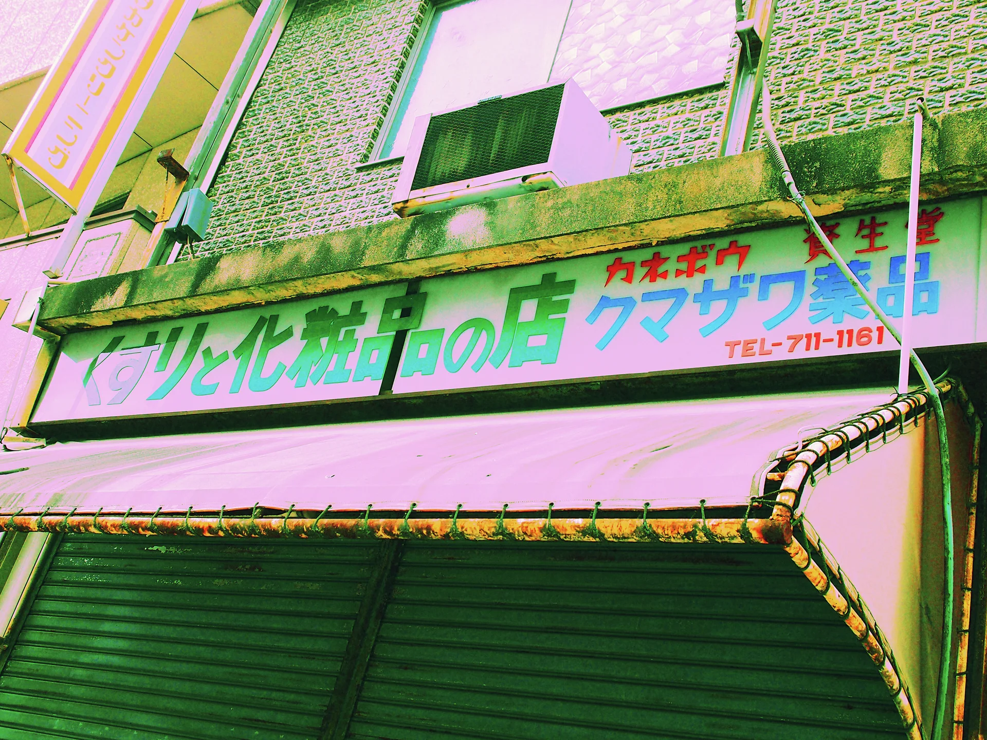 A long Japanese sign hangs above a worn pink awning and closed green shutters on a tiled facade.