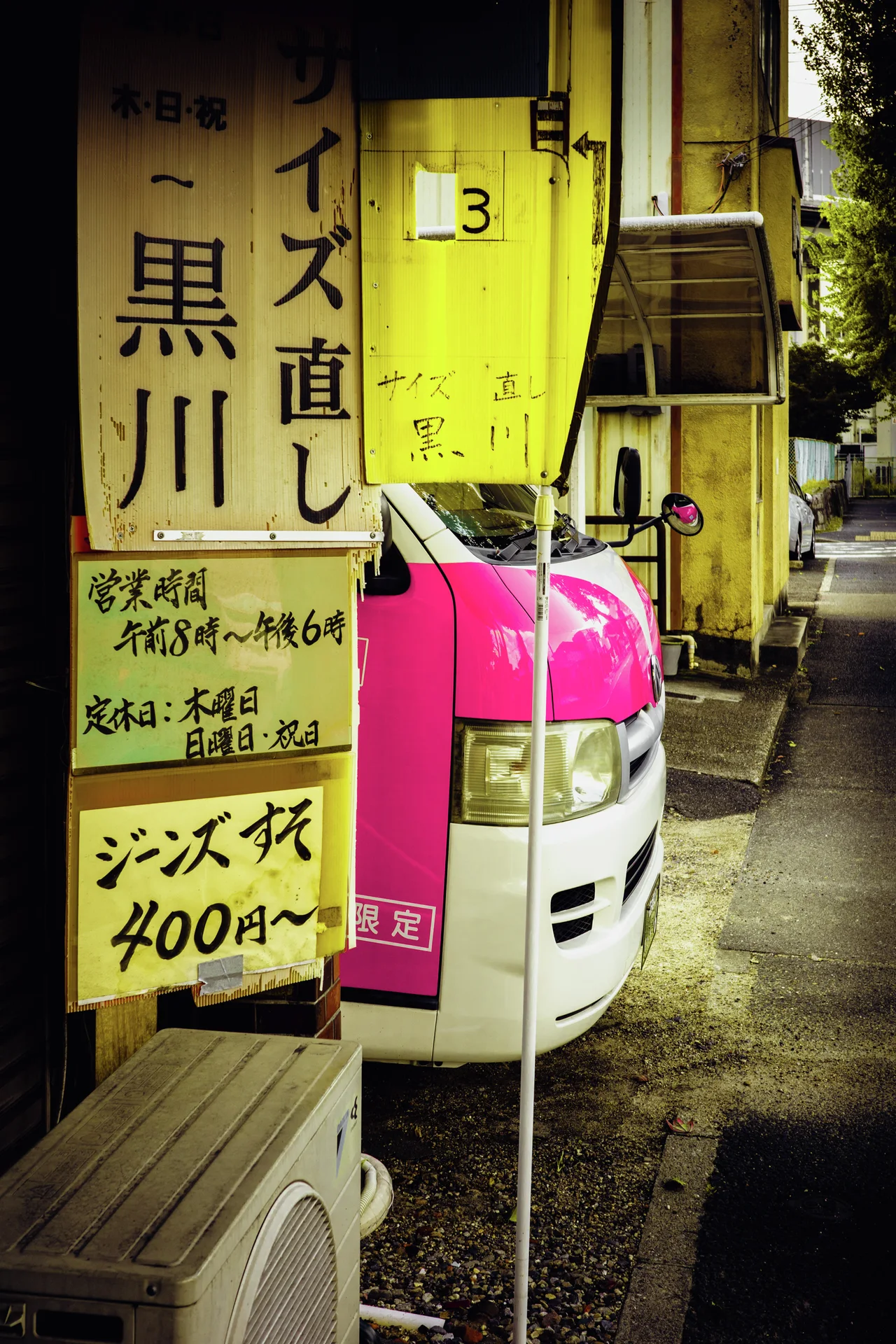 Yellow signs crowd a narrow storefront beside the nose of a pink and white vehicle.
