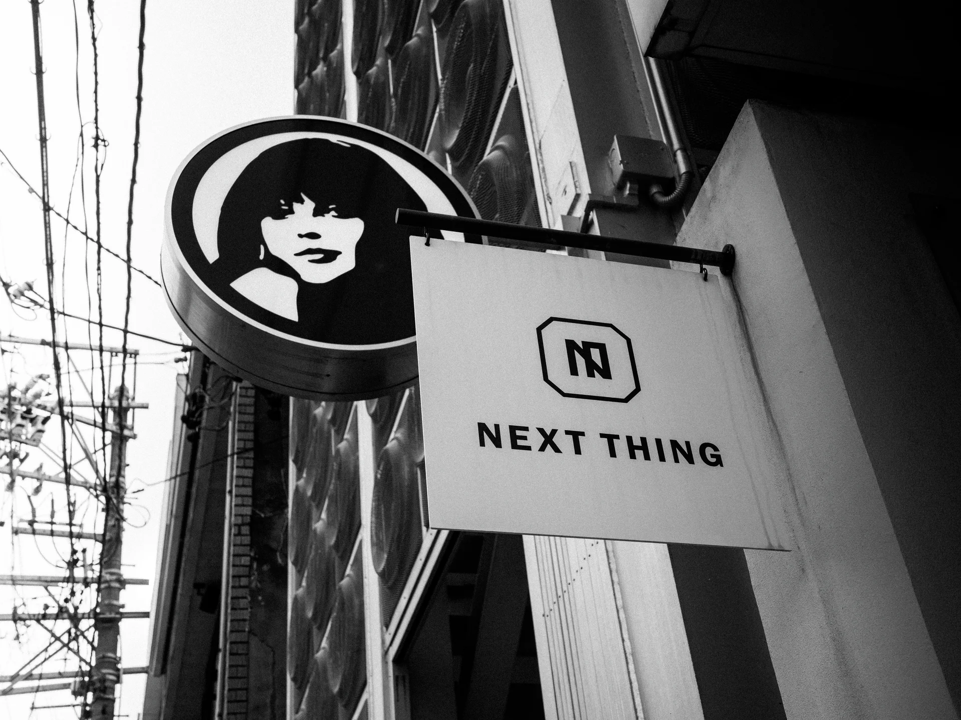 A circular face emblem and pale Next Thing sign project from a building beside overhead wires.