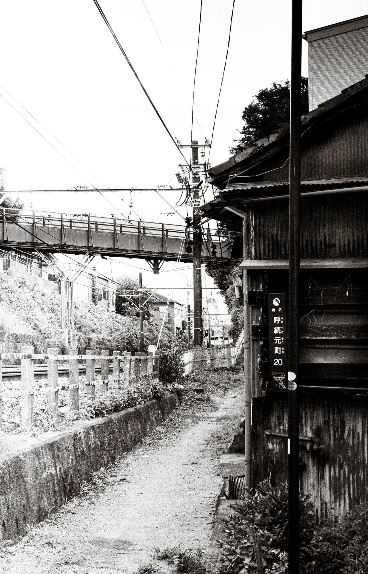 A narrow canal-side path runs beside wooden siding toward an elevated bridge in black and white.