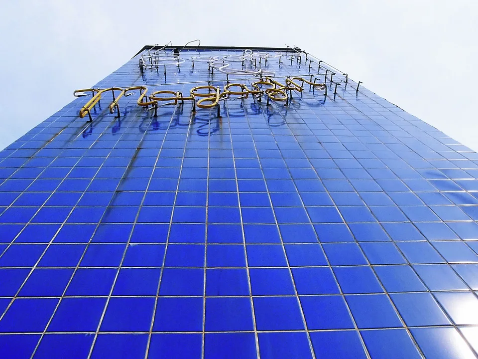 A steep upward view follows a blue tiled wall toward sparse weathered neon lettering against pale sky.