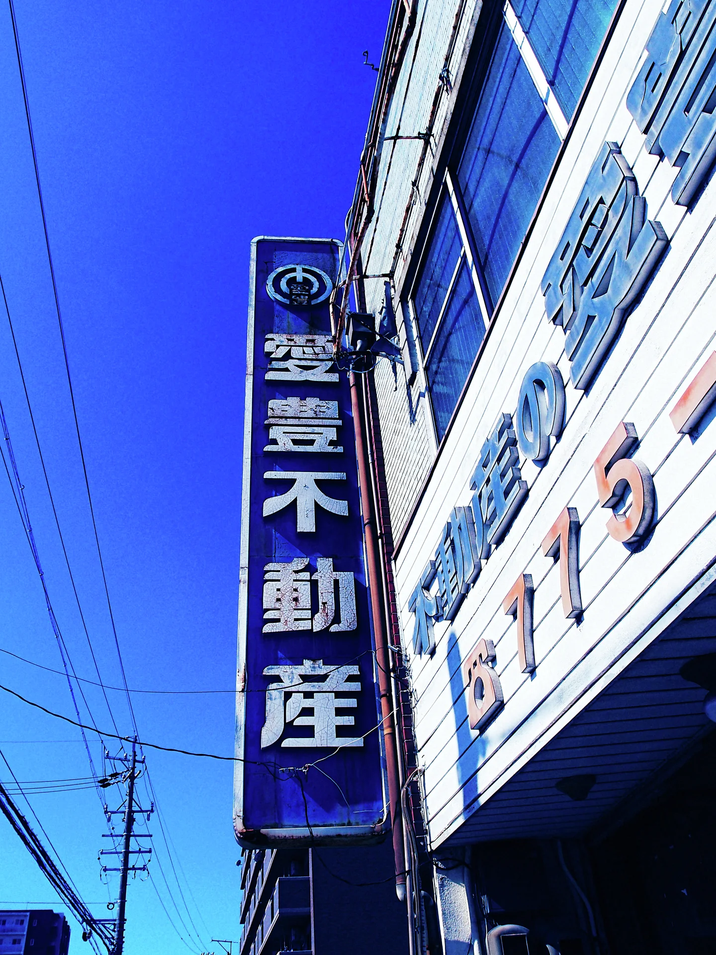 A tall dark vertical real estate sign projects beside bright blue windows under overhead wires.