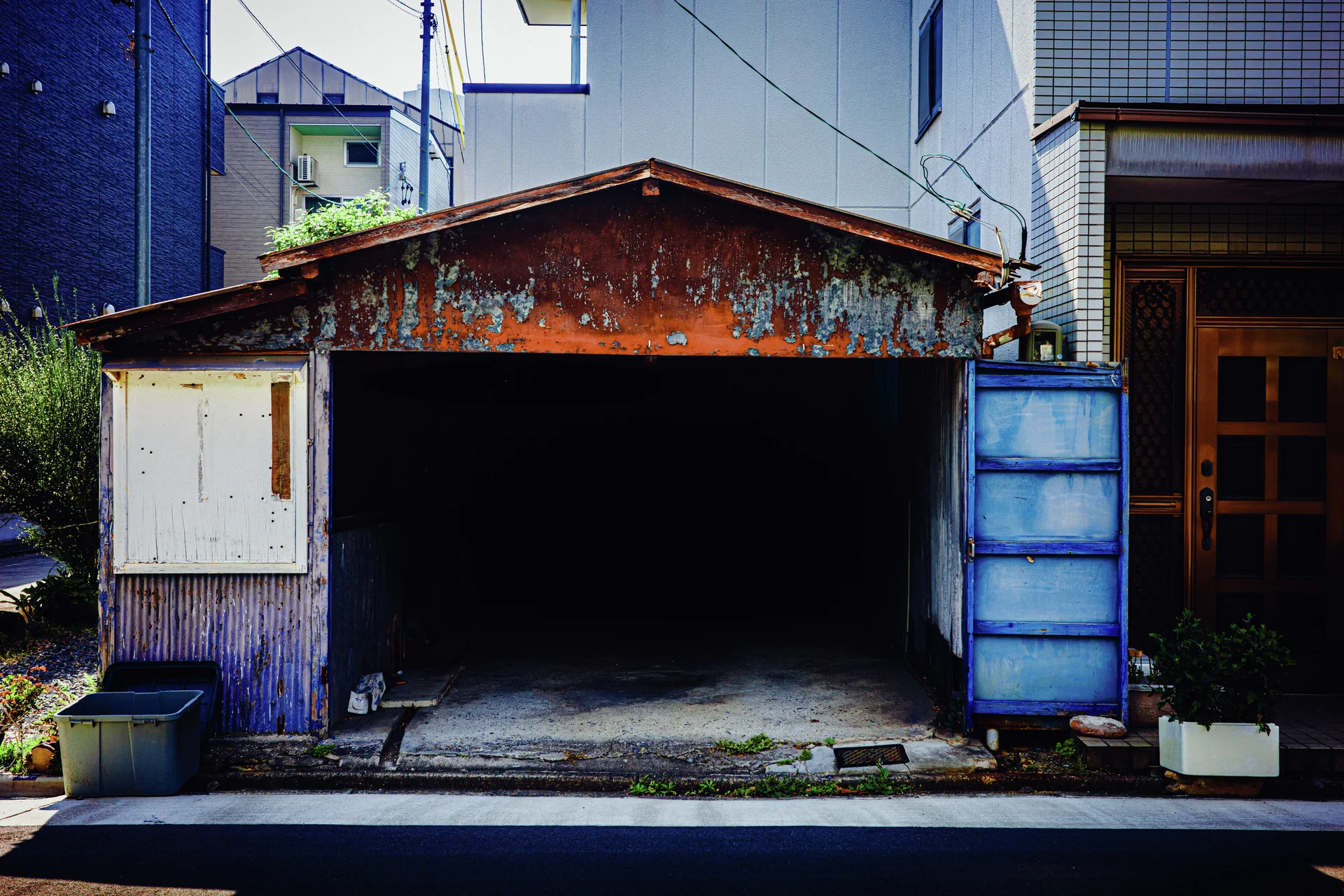 A small open garage has a rust-streaked gabled roof and bright blue door panels beside taller buildings.