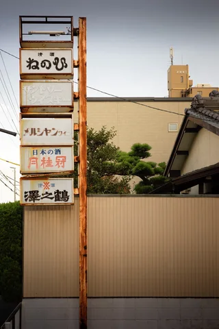 A rusty vertical frame holds stacked rectangular shop signs above a pale fence and nearby rooftops.