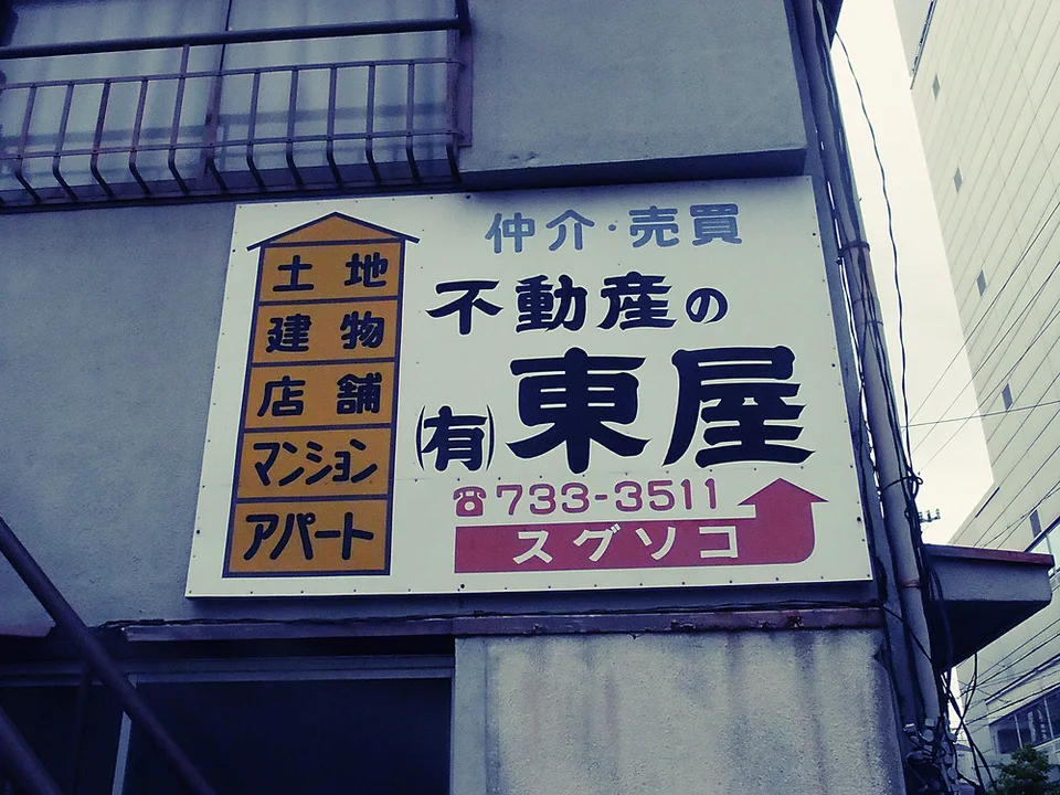 A rectangular real estate sign with black Japanese lettering and a pink arrow hangs below a barred window.
