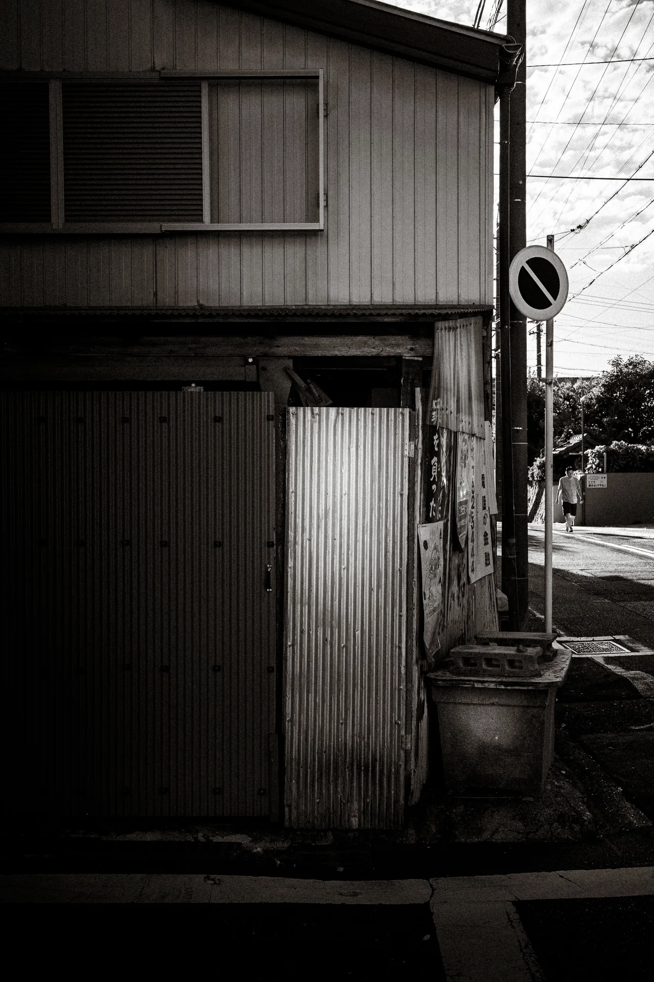 A narrow weathered corrugated house stands beside a roadside sign in black and white.