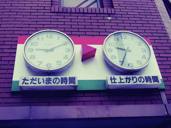 Two round clocks flank a pink arrow on a pale panel mounted against purple brickwork.