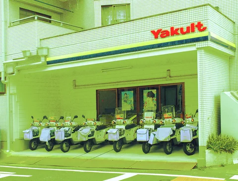 A row of white scooters with front baskets lines a tiled storefront beneath red Yakult lettering.