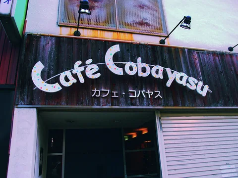 White Cafe Cobayasu lettering decorates a dark wooden sign beneath two angled lamps and upper windows.