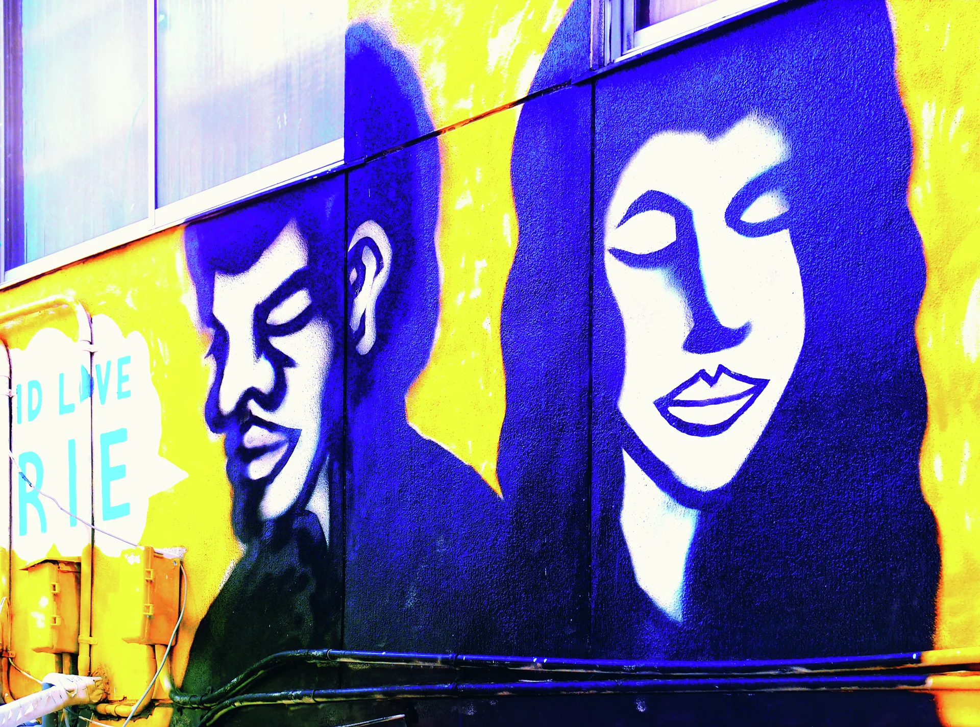 Large stylized blue portraits of a man and woman cover a vivid yellow exterior wall.