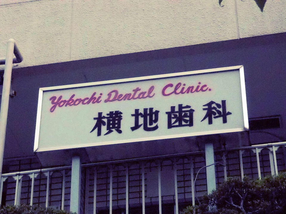 A pale dental clinic sign with pink English script and dark Japanese characters sits above barred windows.