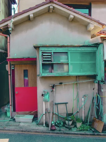 A small pale house has a red door, turquoise sliding window panels, and tools leaning against its wall.