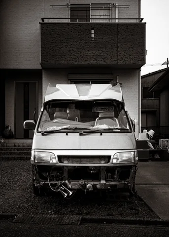 A battered pale van with a crumpled front stands beneath a balcony in black and white.