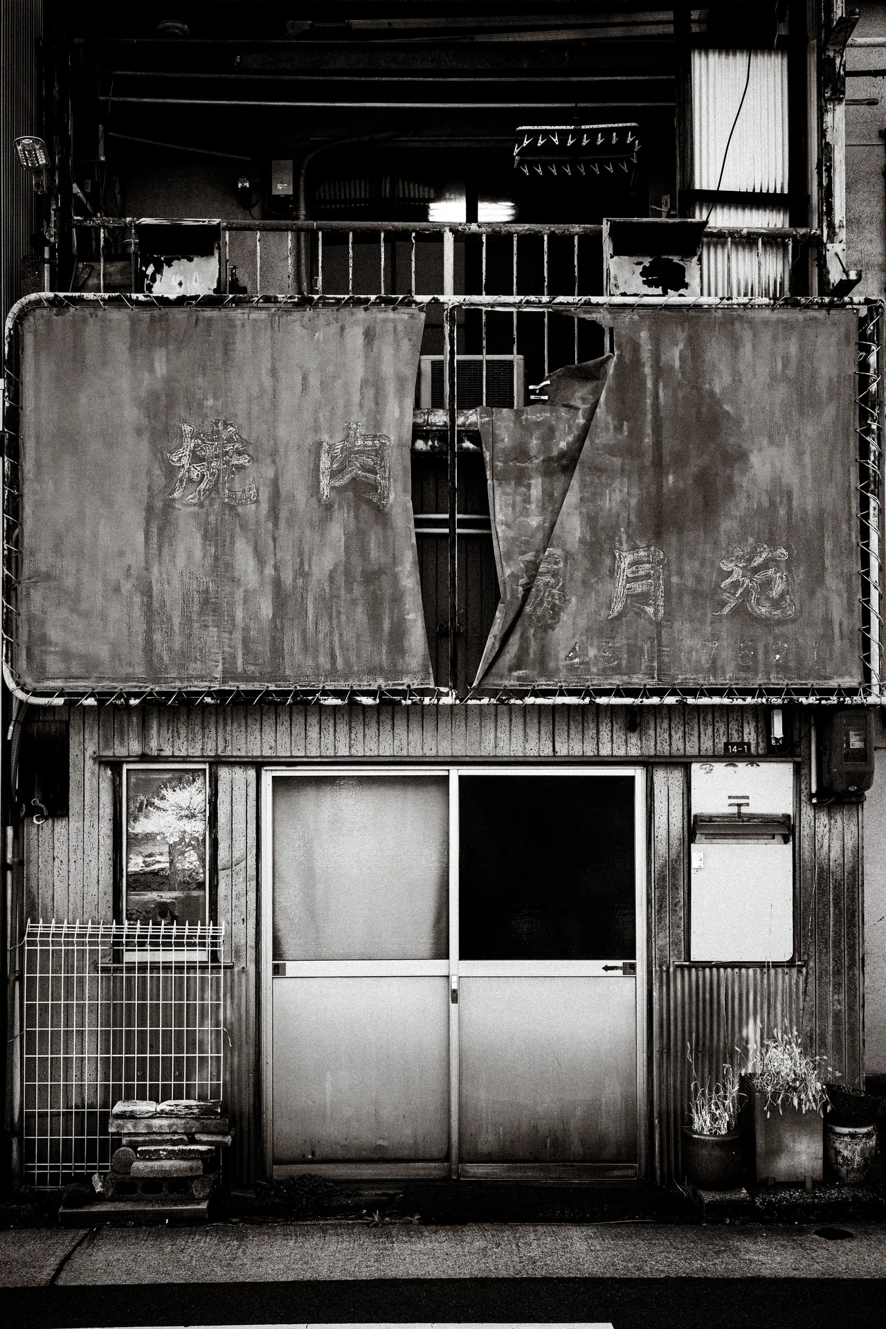 Torn balcony coverings hang above sliding doors on a weathered narrow facade in a black and white view.