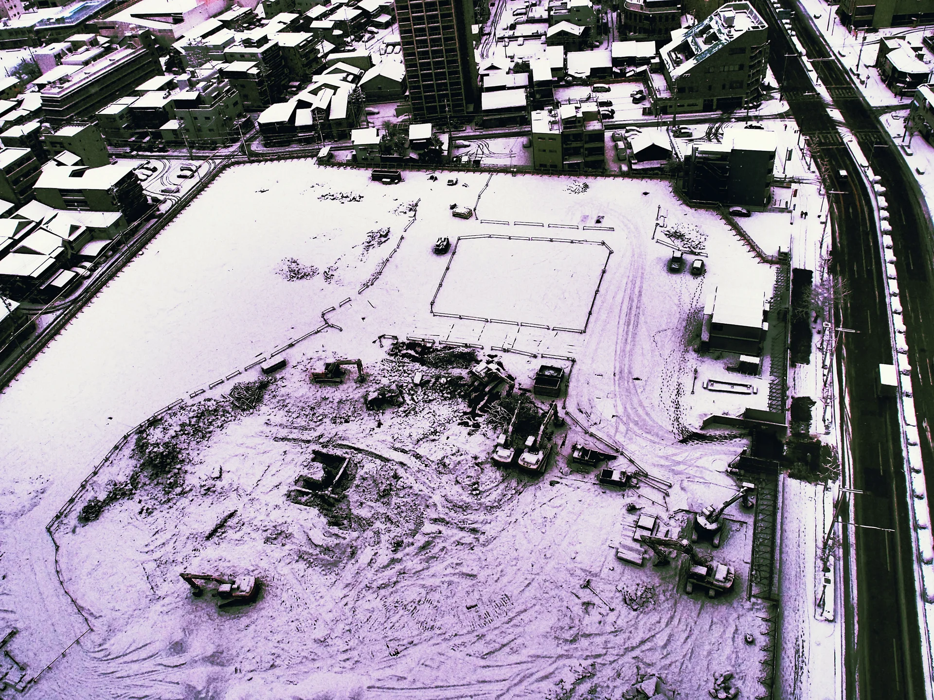 Snow blankets a broad construction site and surrounding rooftops beside a curving canal in black and white.