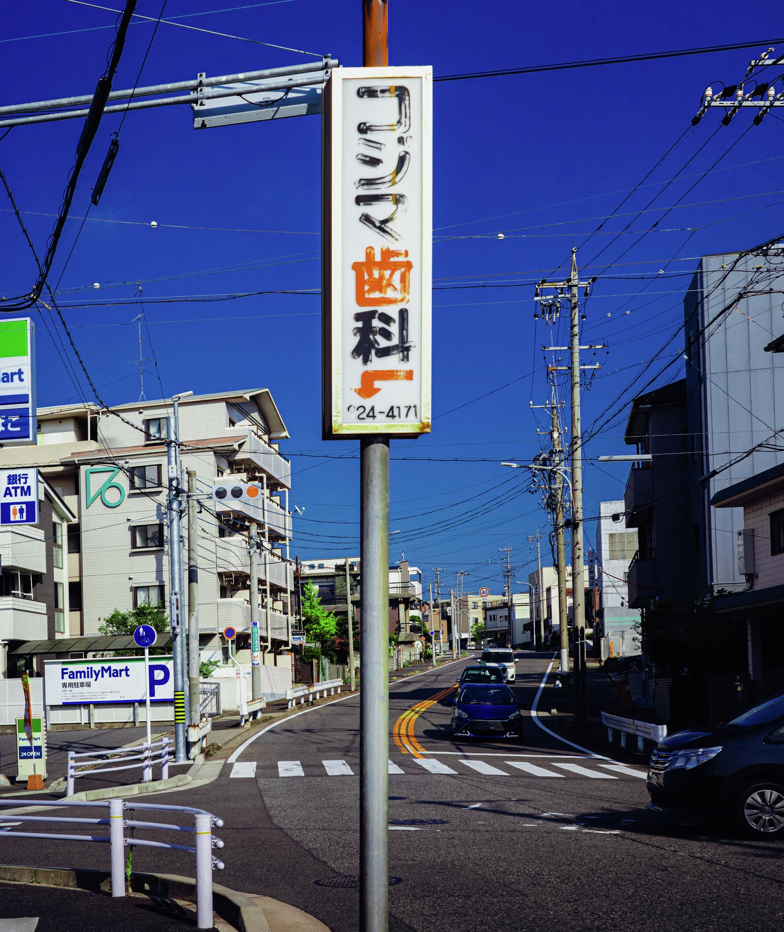 A tall narrow sign rises beside a sunny street intersection crossed by power lines under blue sky.