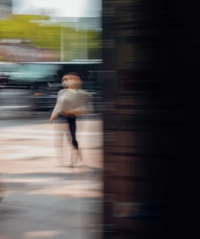 A blurred pedestrian in pale clothing appears beside a broad dark column against streaked street reflections.