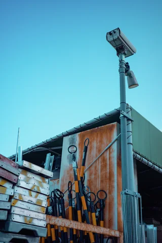 Long-handled tools lean against rusty siding beneath a pole-mounted lamp and a clear turquoise sky.