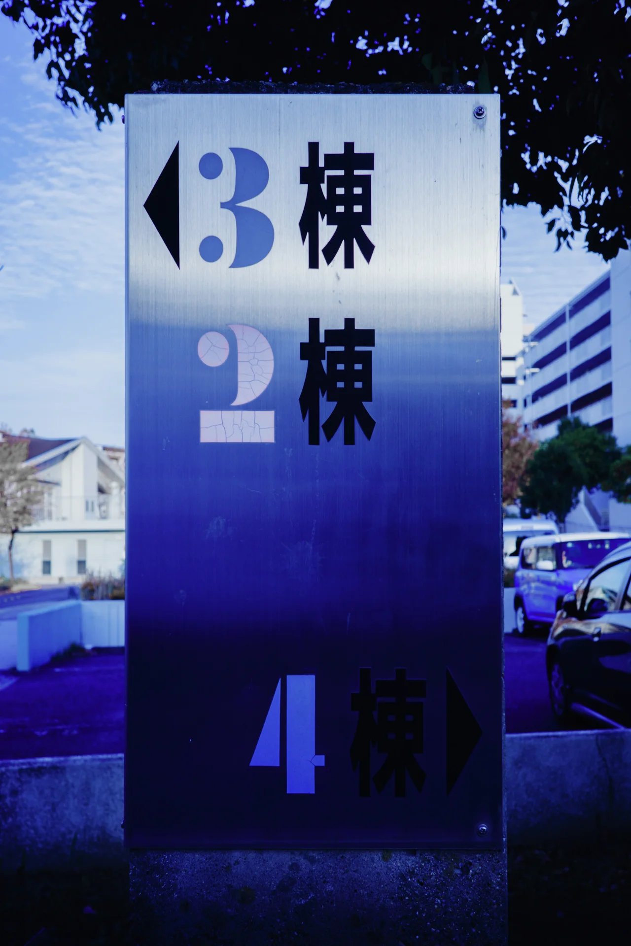 Large pale numbers and dark Japanese characters mark a tall blue-gradient directional sign beneath tree branches.