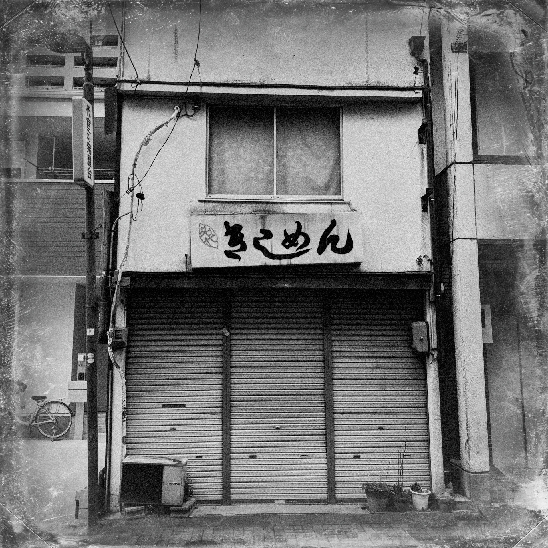 A narrow shuttered shop has Japanese lettering beneath a small upper window in black and white.