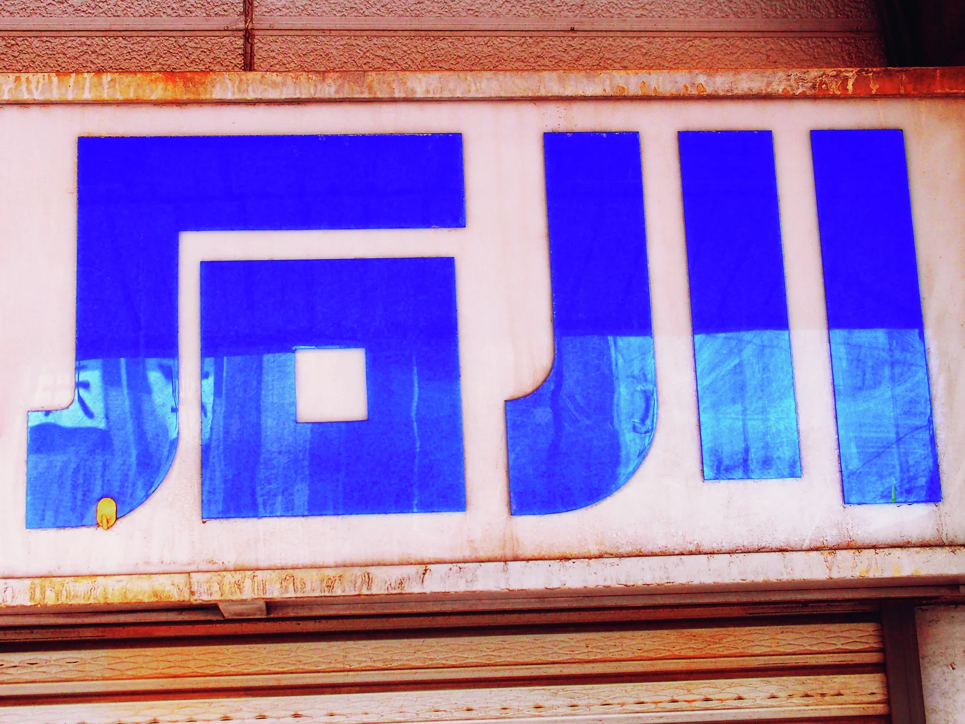Oversized blue geometric lettering fills a weathered pale sign above a metal shutter.