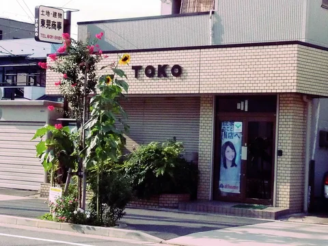 Raised TOKO lettering spans a pale tiled storefront above closed shutters, plants and a portrait-covered door.