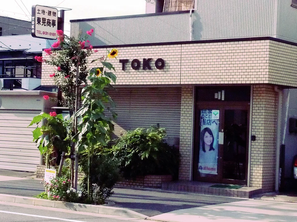 Raised TOKO lettering spans a pale tiled storefront above closed shutters, plants and a portrait-covered door.