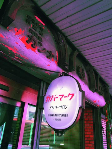 An oval hair salon sign hangs beneath a covered walkway beside a shopfront tinted pink and green.