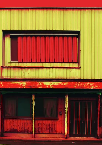 A red shutter sits above curtained windows and a sliding door on a yellow and red building facade.