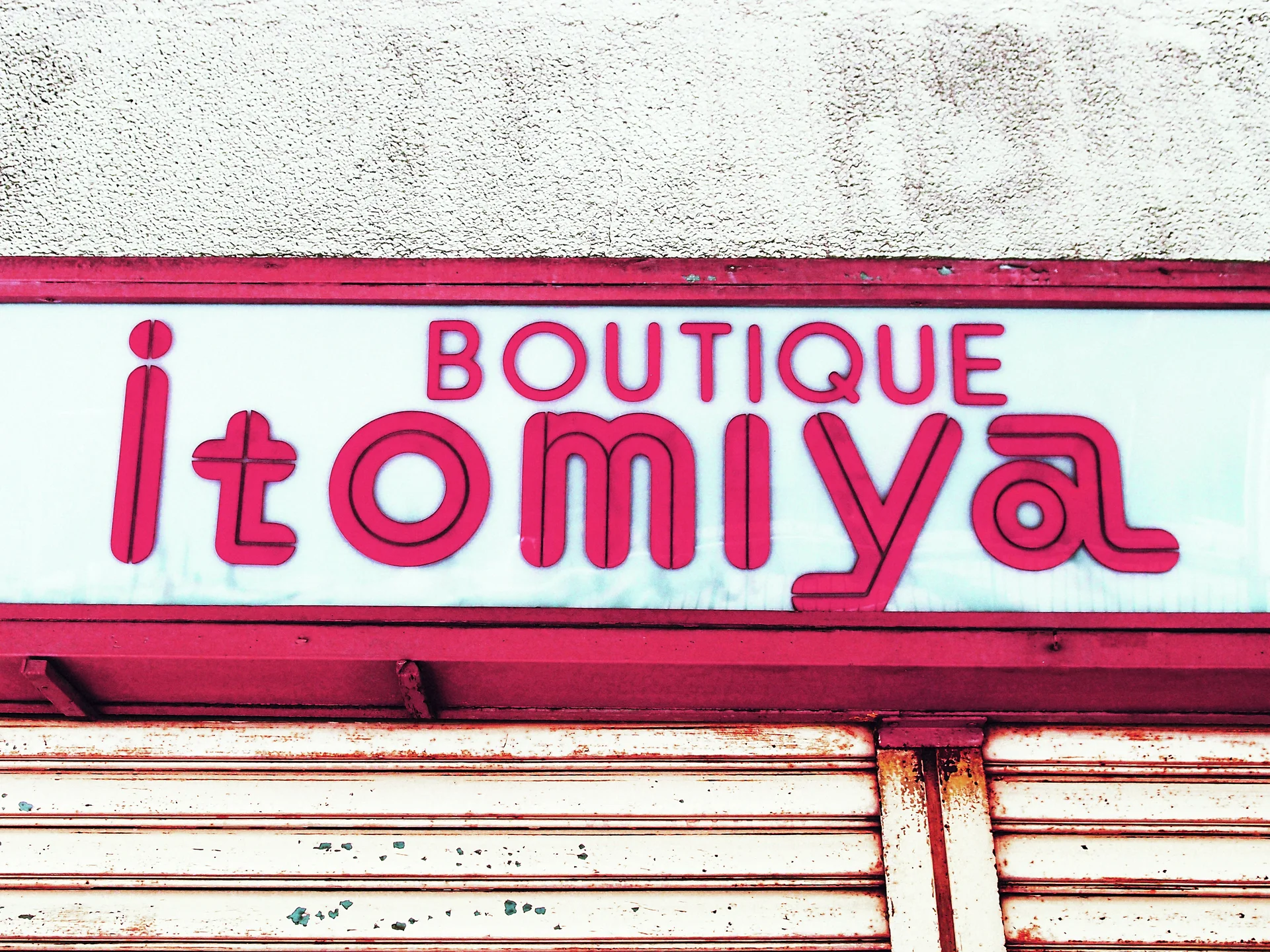 Pink outlined boutique lettering fills a white sign above closed weathered shutters with matching pink trim.