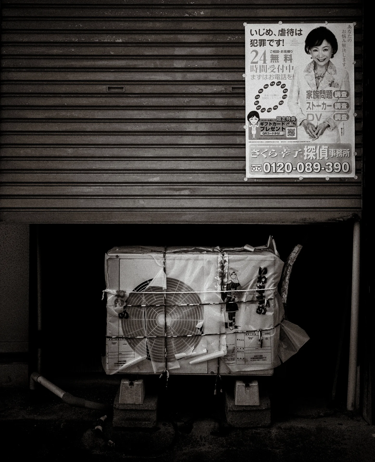 A battered wrapped parcel rests below a closed shutter and portrait poster in black and white.