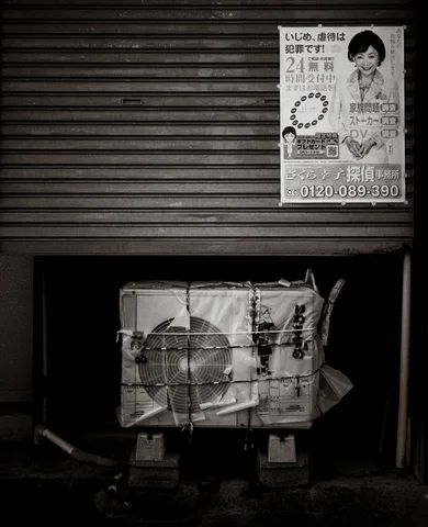 A battered wrapped parcel rests below a closed shutter and portrait poster in black and white.