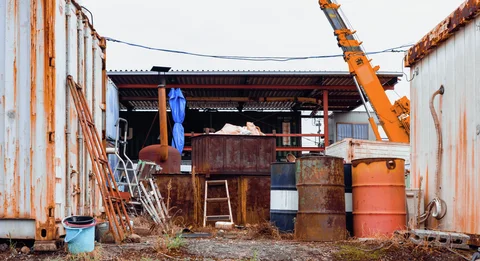 Rusty barrels and leaning ladders crowd a small industrial yard beneath an orange crane boom.