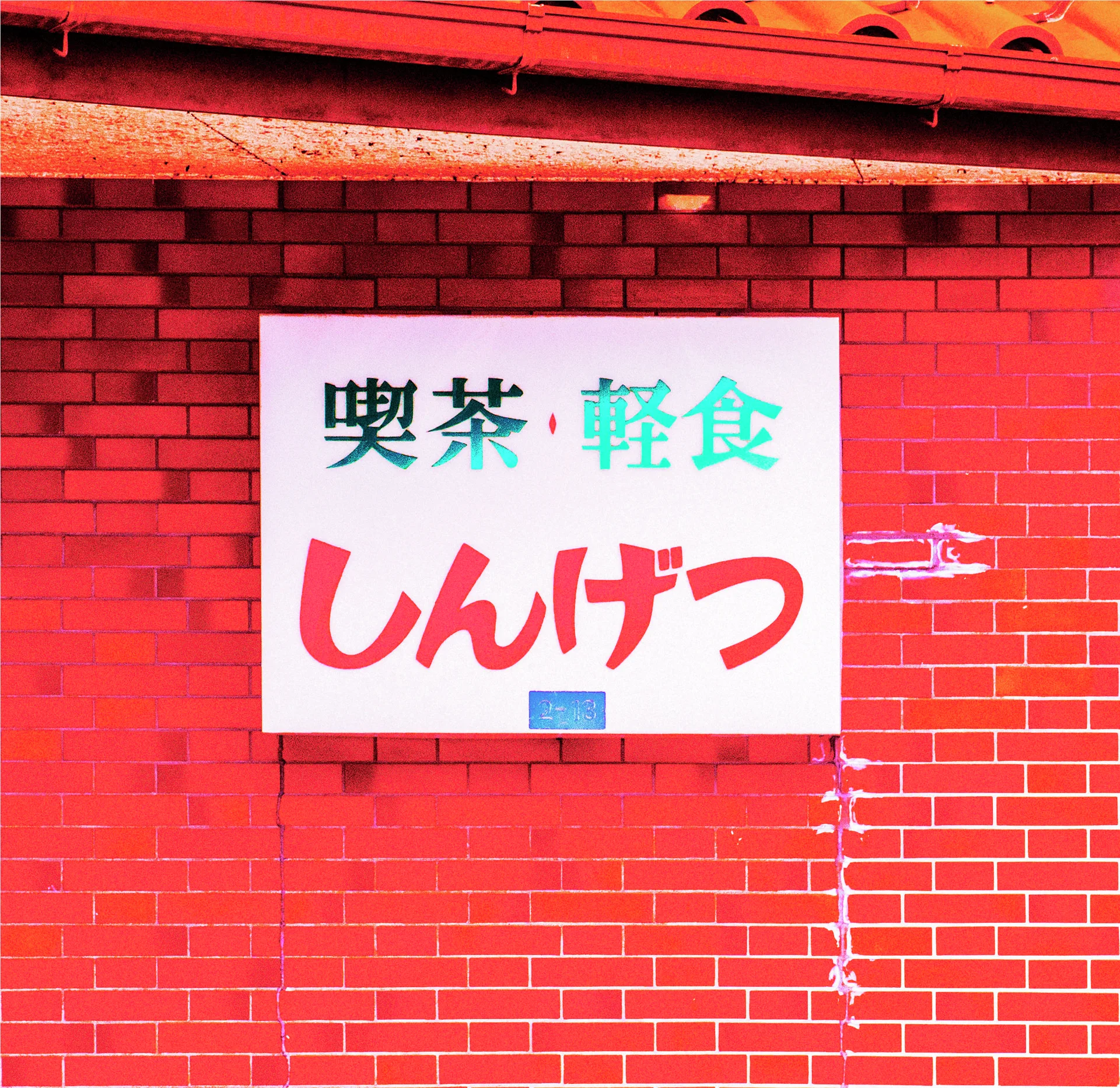 A white rectangular sign with red, black and green Japanese lettering hangs on a red brick wall.
