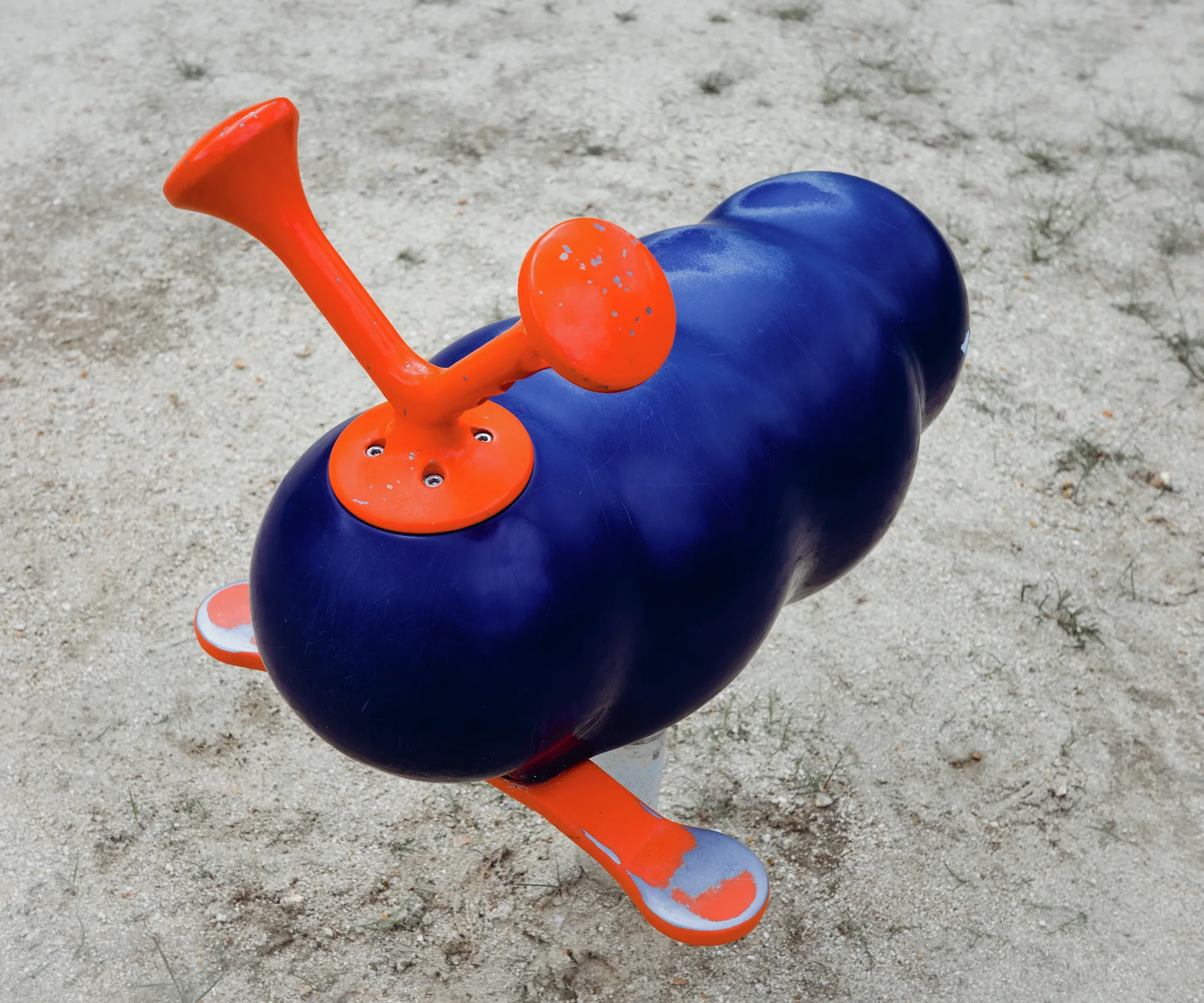 A rounded blue playground rider with orange handles and footrests stands on pale sandy ground.