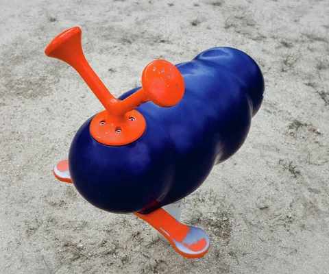 A rounded blue playground rider with orange handles and footrests stands on pale sandy ground.