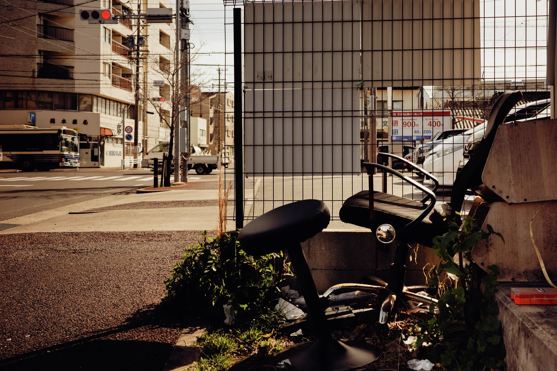 A bicycle and bulky discarded items sit beside a wire fence overlooking a sunlit urban street.