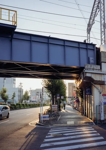 A blue steel railway bridge spans a road and sunlit sidewalk beside shuttered storefronts.