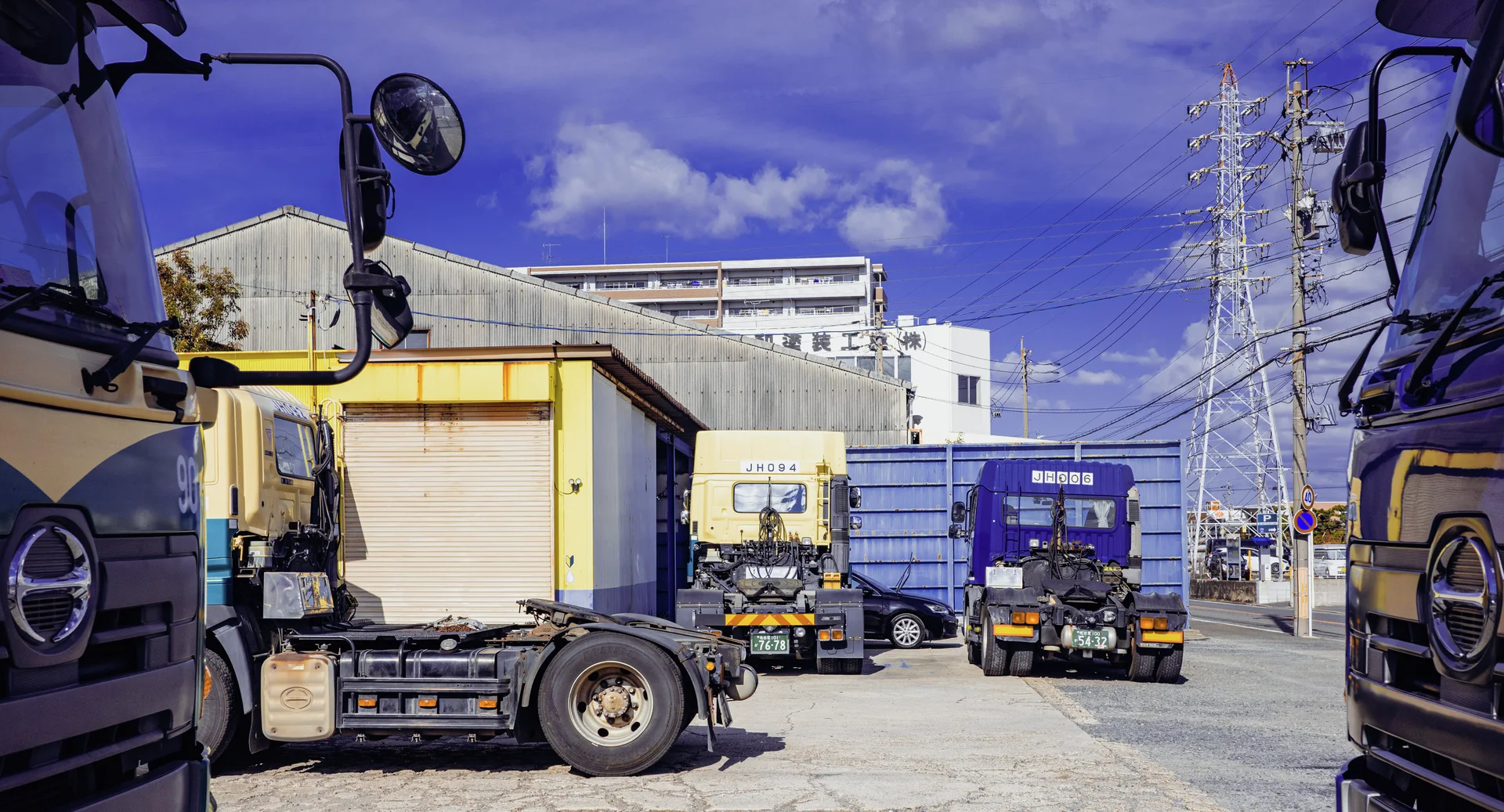 Parked trucks frame an industrial yard with a yellow-trimmed building and distant electricity pylons beneath blue clouds.