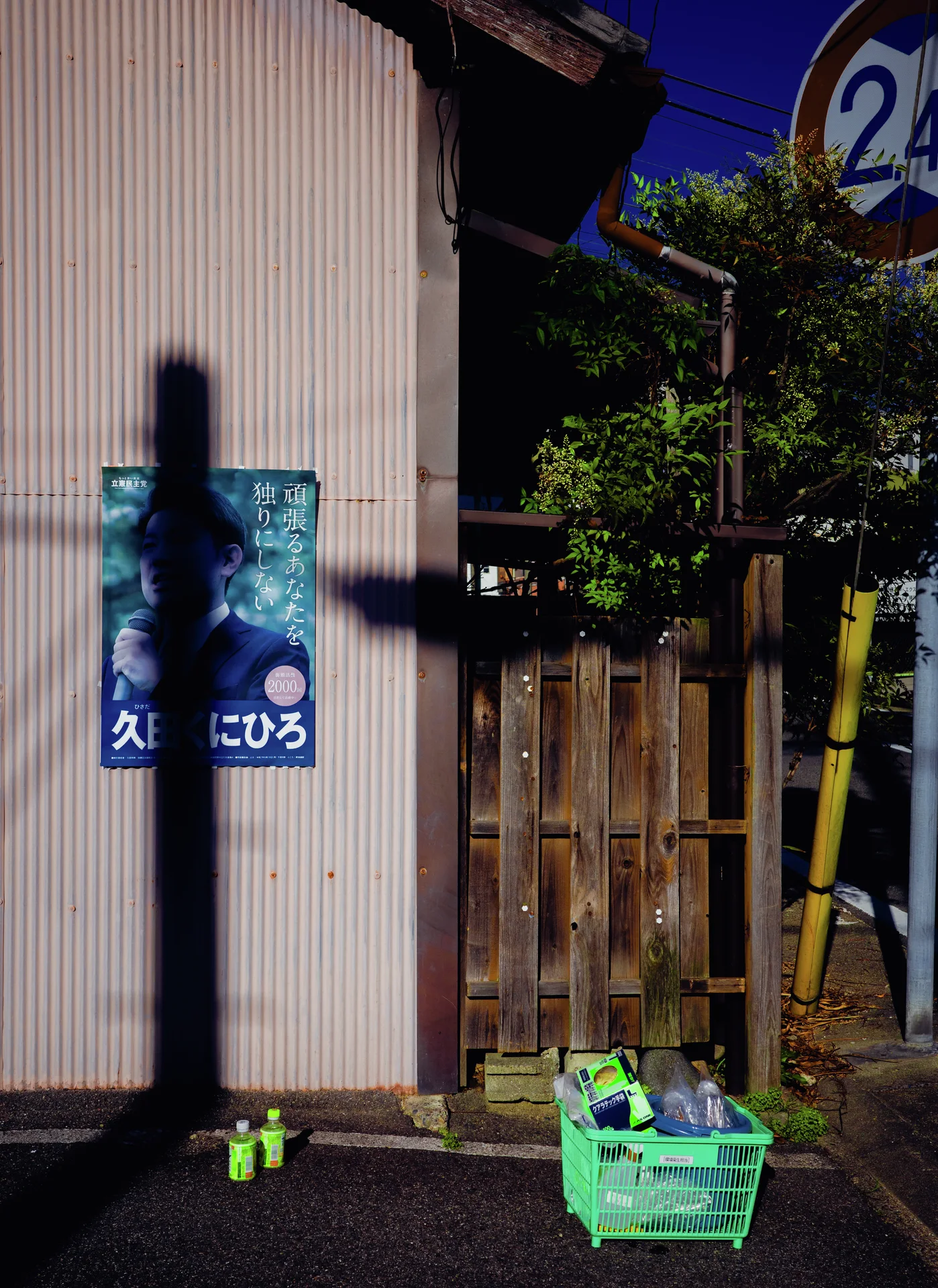 A pole shadow crosses a portrait poster above green crates outside a corrugated building.