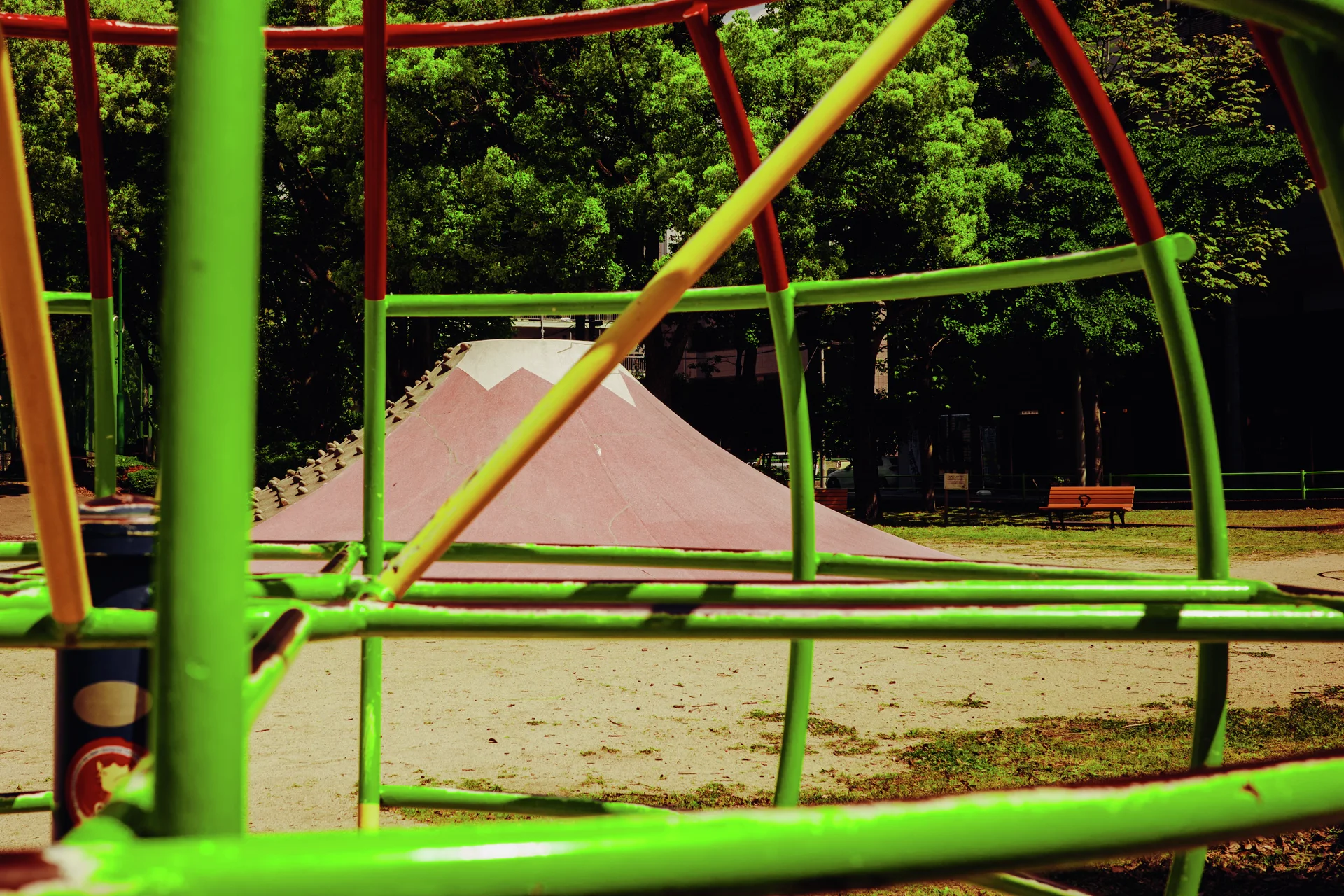 Green and yellow playground bars frame a pink mound with trees beyond a sandy ground area.