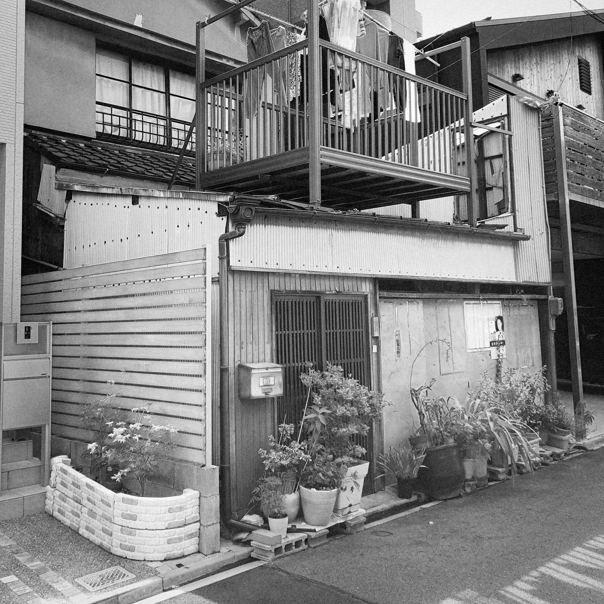 Potted plants line a small weathered residence beneath a raised metal balcony in black and white.